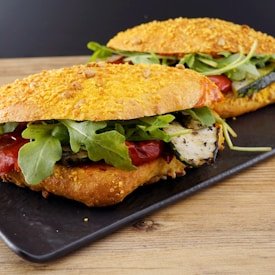 Two gourmet chicken sandwiches with arugula, tomatoes, and grilled bread on a black tray