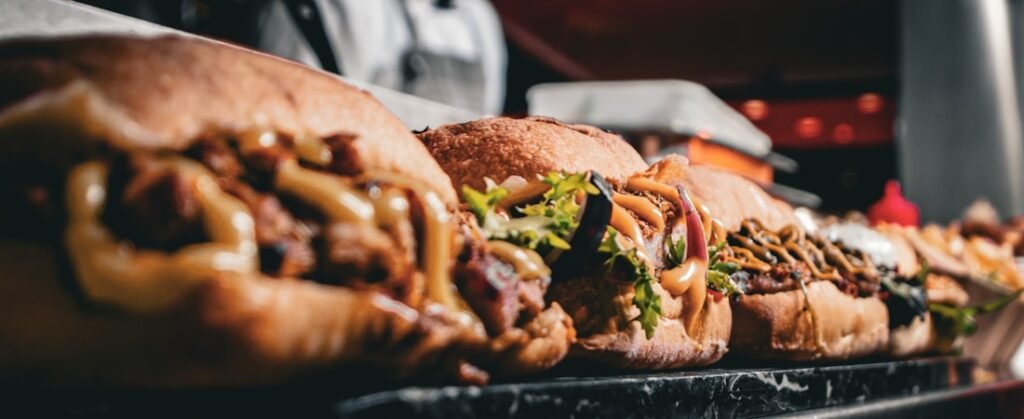 Row of loaded sub sandwiches topped with melted cheese, greens, onions, and sauces